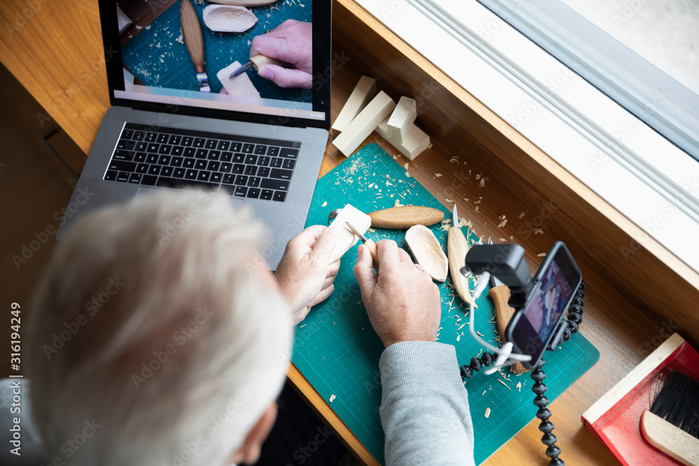 An older adult's hands are seen as he carves a block of wood. A camera is capturing his work and displaying it on a laptop screen.